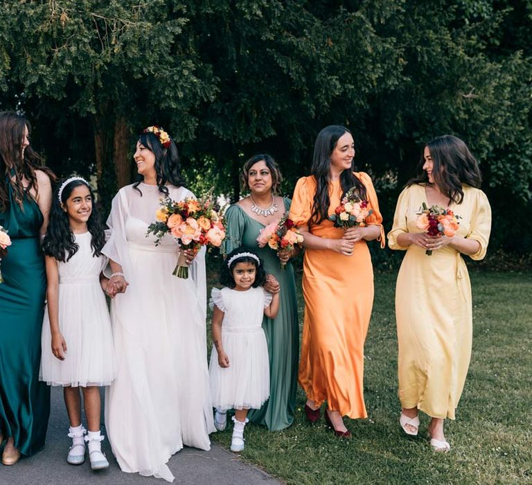 Bride in long sleeve mesh overlay wedding dress and colourful bridal flower crown walking with bridal party in mismatched bridesmaid dresses and flower girls in white tulle flower girl outfits 