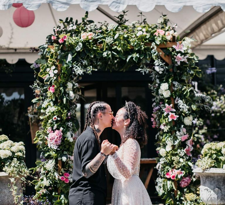 Bride in long sleeve v-neck sheer polka dot overlay sparkly wedding dress and gold celestial bridal headband kissing bride in black shirt and black trousers at Viking themed wedding