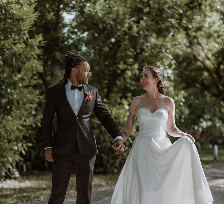 Bride in strapless sweetheart neckline wedding dress walking hand in hand with groom in classic black tuxedo with bowtie and pink and orange wedding boutonniere at The Old Rectory Berkshire wedding venue