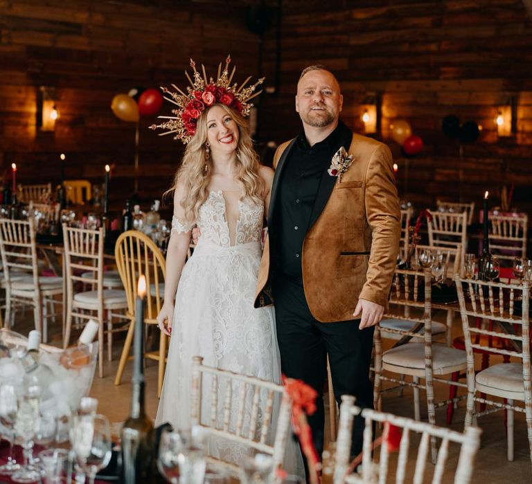 Bride in off shoulder lace wedding dress with corset style top and red rose and gold halo bridal crown standing with groom in crushed velvet burnt orange grooms blazer and dried flower boutonniere in Southlands Barn wedding venue reception room with gothic rock and roll wedding tablescape with deep red wedding table runner, black plates and black napkins