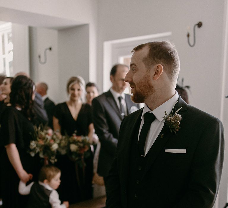 The groom in a classic black wedding suit turns to look at the bride walking down the aisle 