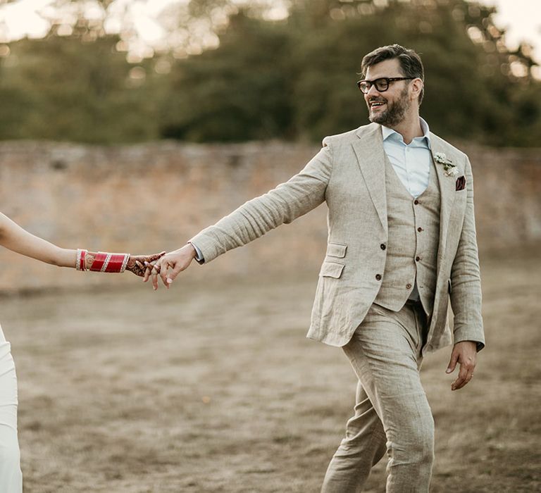 Groom in three piece cream wedding suit walking along with the bride in a fitted slip dress 