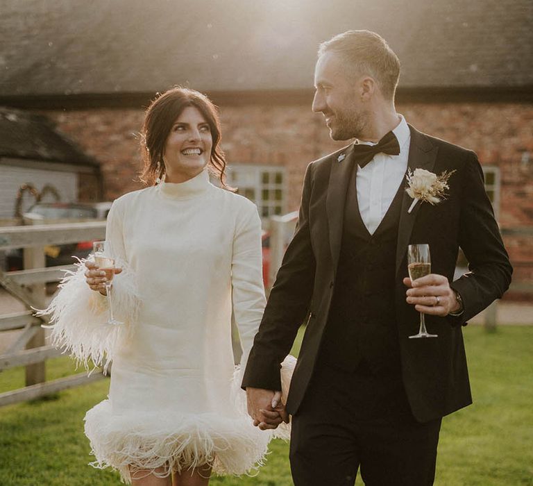 Bride in short feather wedding dress walking with groom in black tuxedo for their couple portraits at golden hour