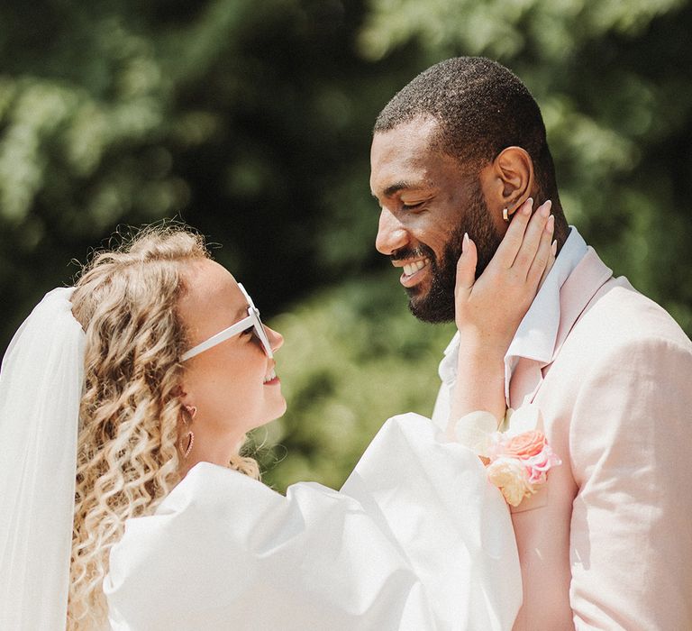 Bride in Watters puff sleeve statement wedding dress with veil and white retro sunglasses smiling up at the groom in a pink suit 