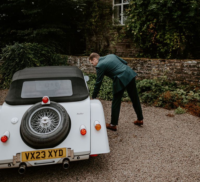 Groom in green suit getting into white vintage wedding car 