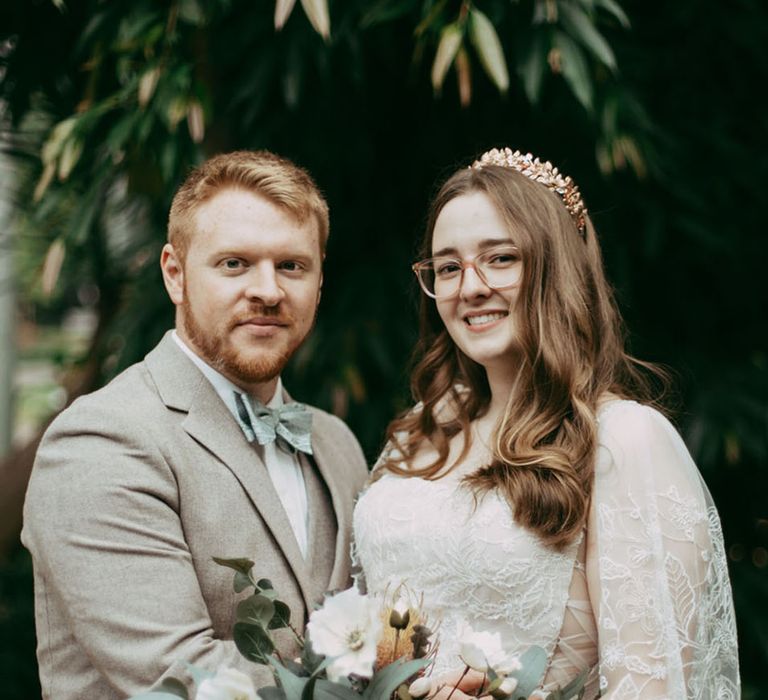 Bride in embroidered wedding dress and cape holding white flower wedding bouquet with the groom in a grey suit with green bow tie 