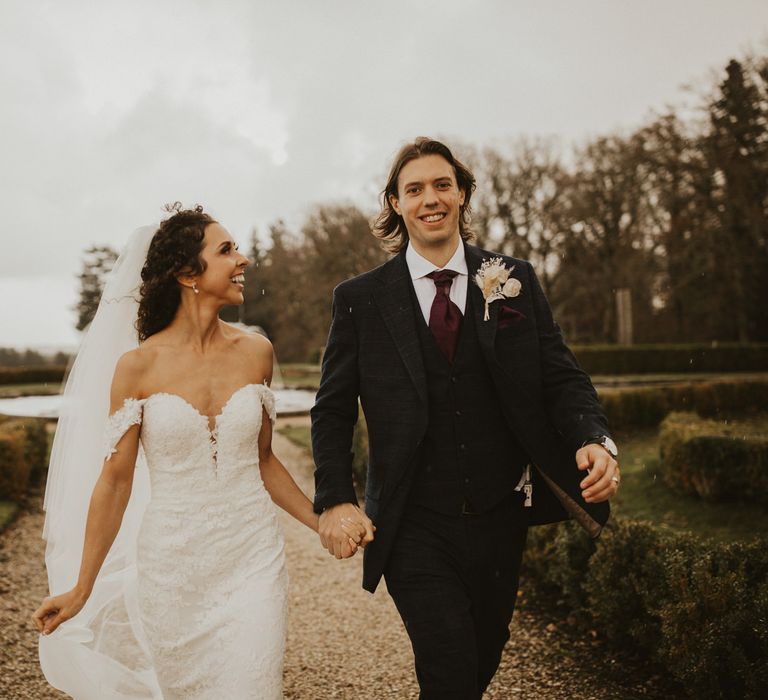 Wedding portrait of a bride in an off the shoulder lace dress holding hands with her groom in a navy suit and burgundy tie at Rhinefield House wedding venue