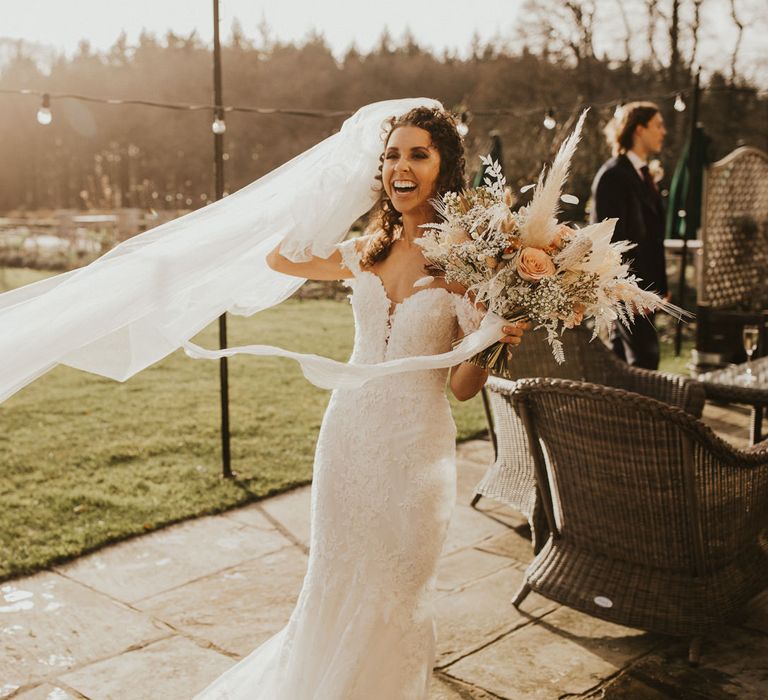 Bride with brown curly hair holding onto her cathedral length veil in a lace off the shoulder wedding dress