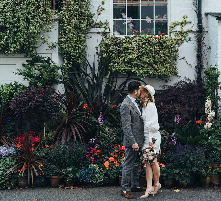 Bride and groom kissing in front of garden florals on the streets of London, bride wears a short wedding dress and hat