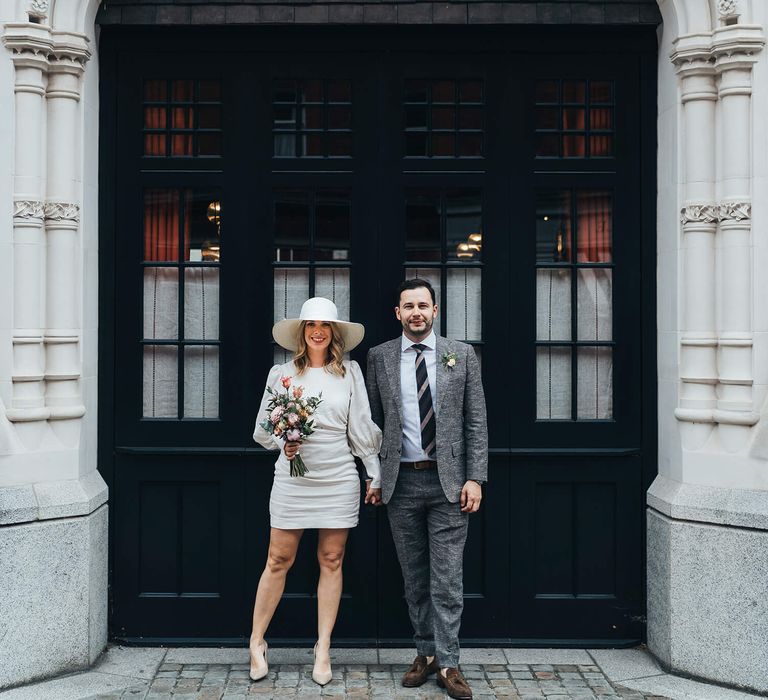 Bride and groom wedding day photography under arched door way, bride wearing short Net A Porter wedding dress with pink and coral bouquet groom  wearing grey suit