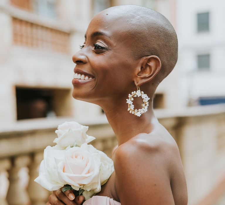 Black bride in a pink chiffon off the shoulder bridesmaid dress and jewel earrings holding a posy of white roses, laughing 