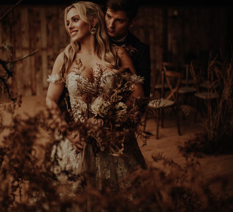 Groom embracing his bride in an off the shoulder lace dress as she holds a dried flower bouquet 