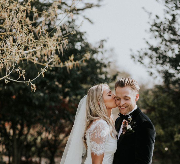 Bride in white Charlie Brear dress, white lace Augusta Jones top and full length veil kissing groom in black velvet Hugo Boss suit in the grounds at Caswell House wedding