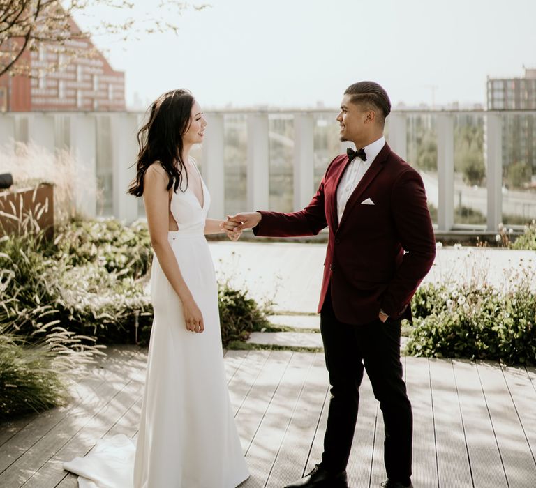 Stylish groom in a dark red jacket admiring his bride in a minimalist wedding dress at their first look.