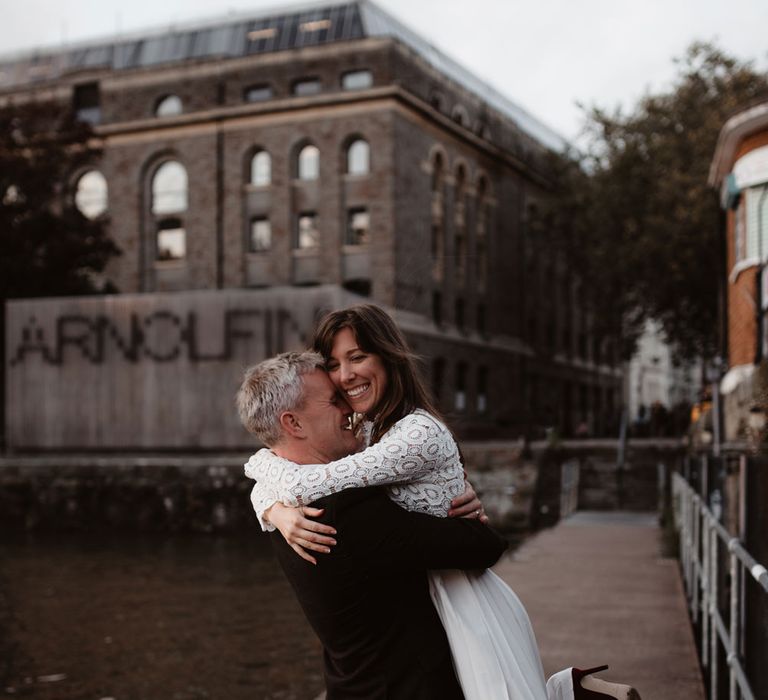 Bride in red suede heels and long sleeved lace top Self Portrait dress with pleated skirt dances with groom in brown woollen blazer and grey trousers on dock at Bristol 