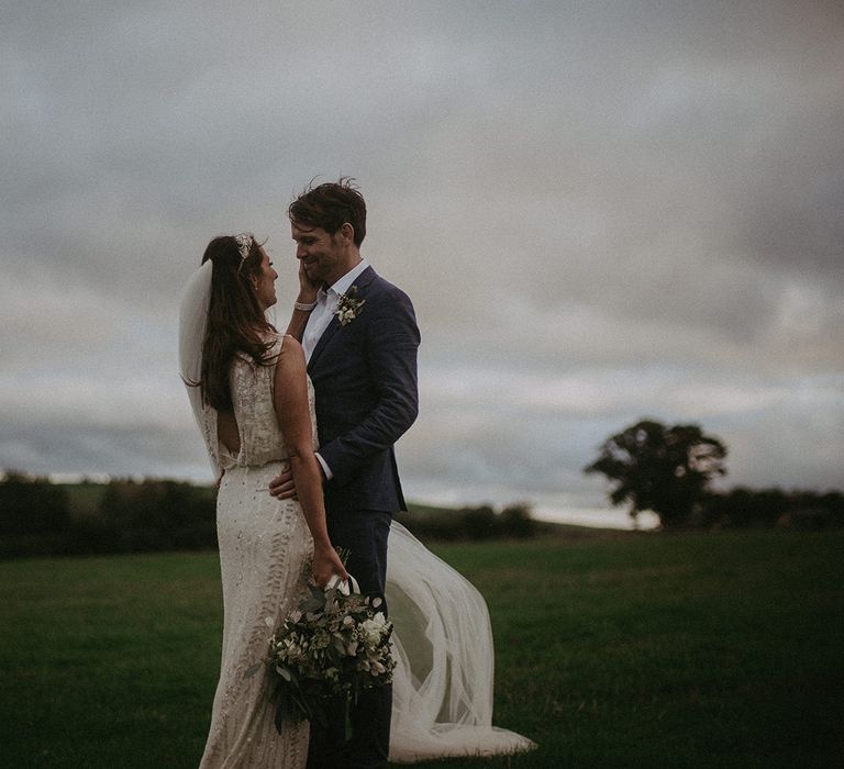 Bride & groom stand together outdoors in green fields on their wedding day