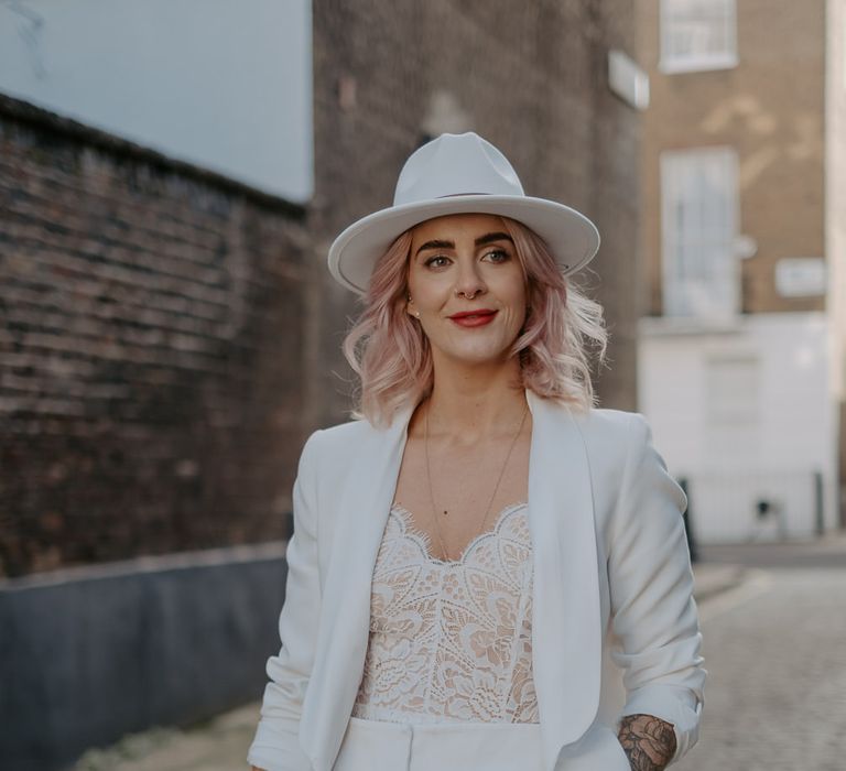 White bride in white suit and fedora hat holding bouquet and other hand in pocket