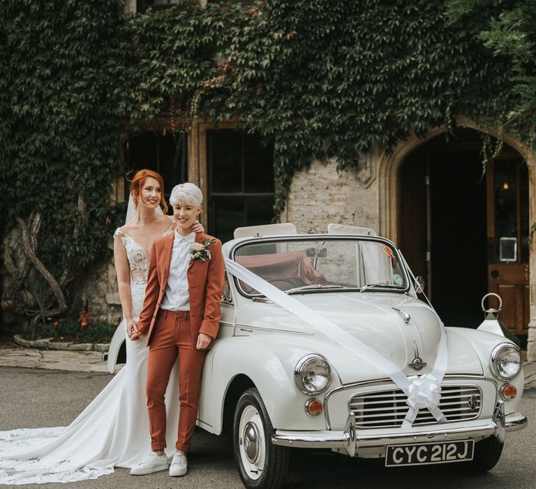 A same-sex couple stand in front of a classic white wedding car. The woman in front wears a terracotta suit and has platinum cropped hair. The woman behind wears a white wedding dress and has her red hair loosely tied back. Photography by Steph Dreams Photos. 