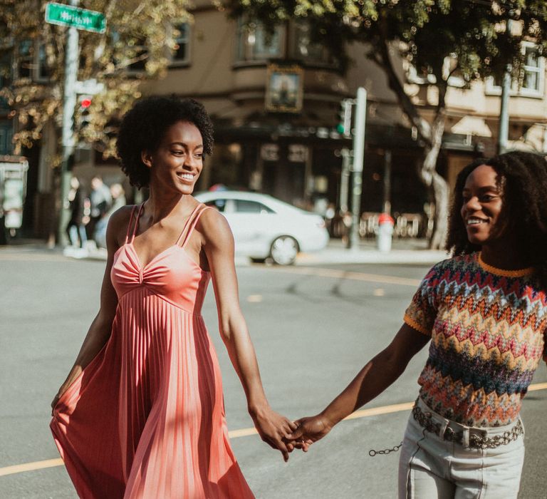 Bride-to-be in a coral dress holding hands with her bride-to-be in a patterned knit crossing the road in San Francisco 