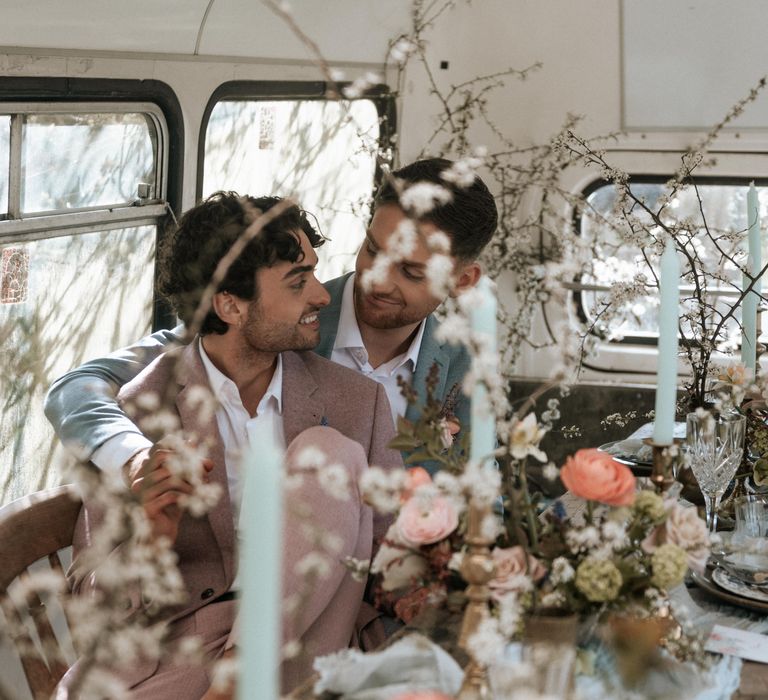 Grooms sit amongst rustic floral displays on their wedding day whilst looking lovingly at one another