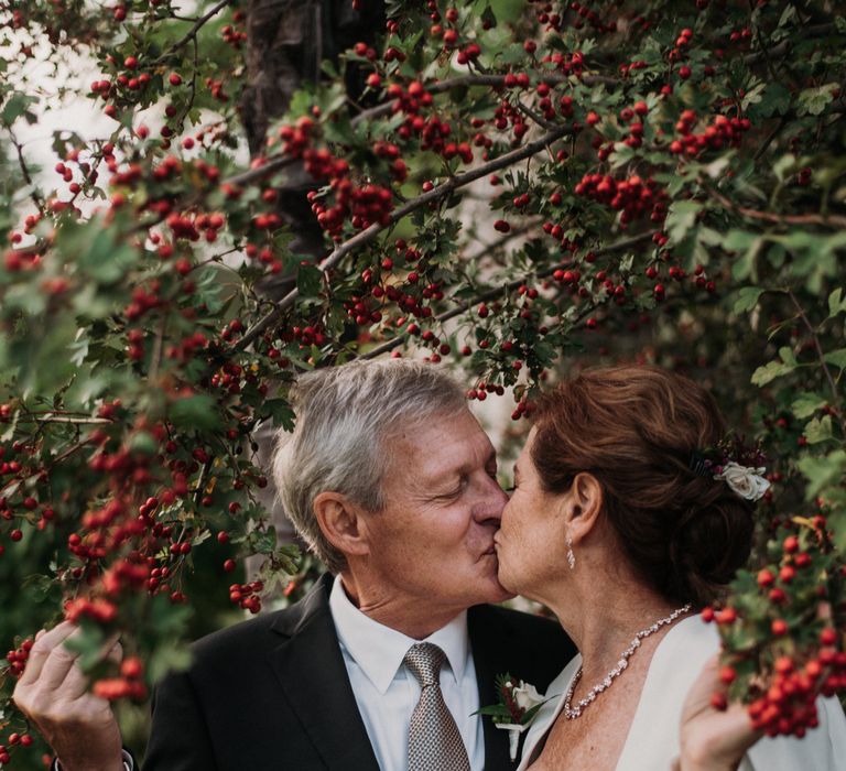 Bride & groom kiss beneath red berry tree whilst holding the branches