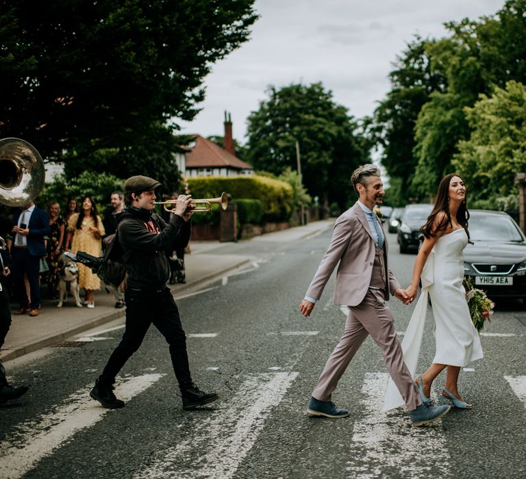 Bride in white strapless Rebecca Vallance Dress and blue slingback heels carrying bridal bouquet walks across zebra crossing holding hands with groom in brown Moss Bros suit and blue tie as they're followed by the New York Brass Band and wedding guests after Harrogate wedding