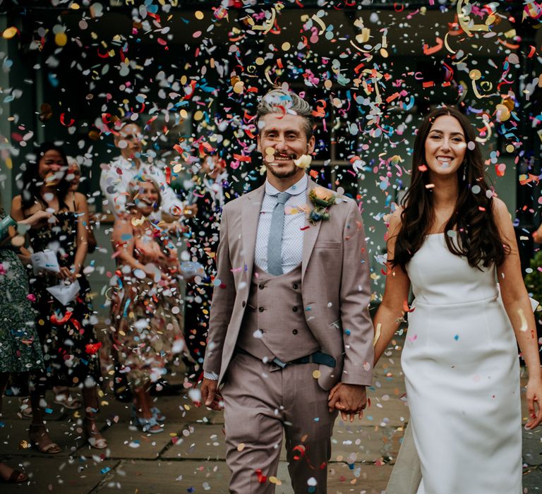 Bride in white strapless Rebecca Vallance Dress holds hands with groom in brown Moss Bros suit and blue tie as they walk through multicoloured confetti at the Hotel du Vin after Harrogate wedding