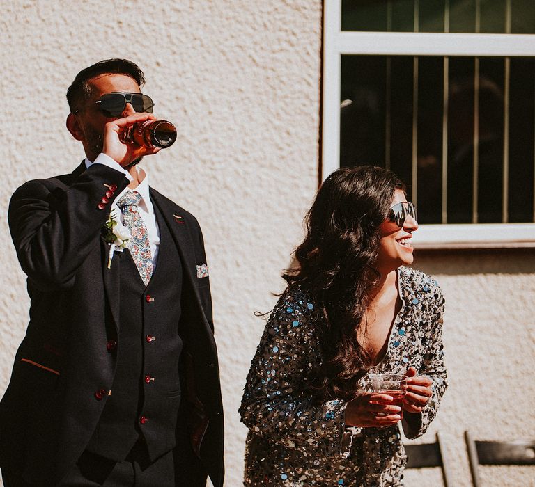 Groom in a three-piece suit with floral tie drinking beer at his garden party wedding reception 