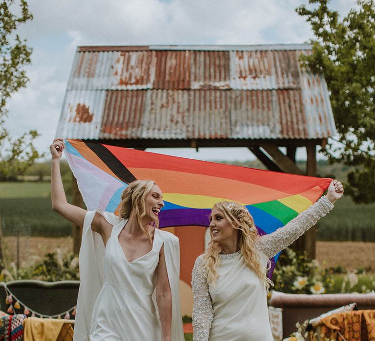 LGBTQI+ couple in a short wedding dress and bridal jumpsuit holding a colourful Pride flag 