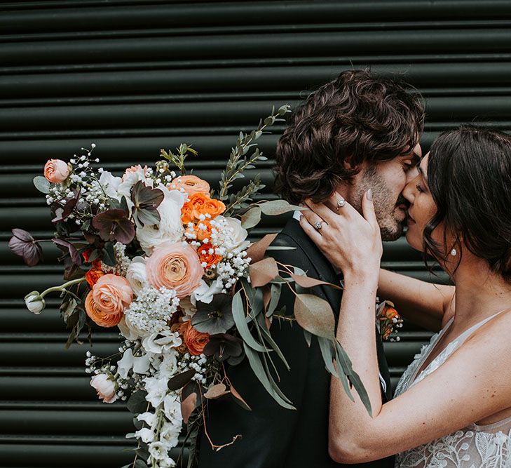 Bride kissing her husband holding her orange, white and green wedding bouquet with ranunculus, spray and avalanche roses and foliage over his shoulder 