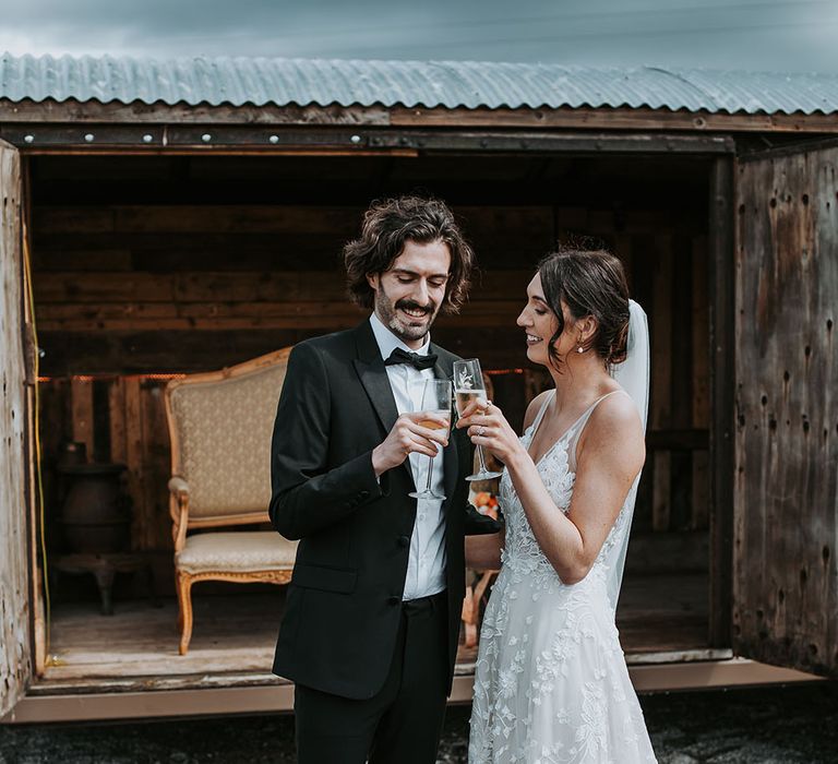 Bride in an appliqué wedding dress with cathedral length veil toasting with her husband in a black tuxedo in the courtyard at Gyhll Barn 