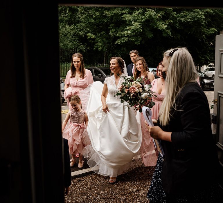 Bride in lace top cap sleeved wedding dress holding white, pink and green bridal bouquet has skirts held up bu bridesmaids in pink dress as she walks into wedding ceremony at The West Mill Derby