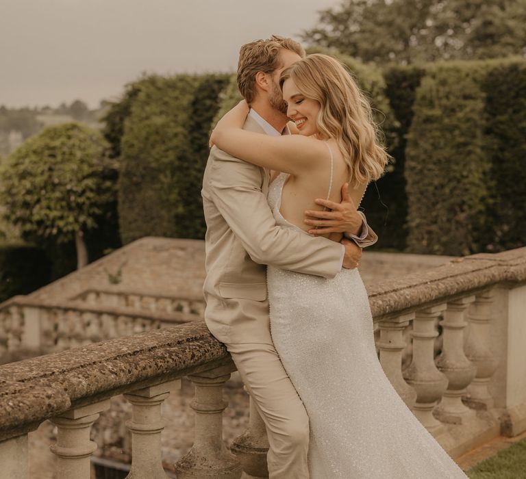 Groom in a beige suit and tan brogues embracing his bride in a sparkly Made With Love wedding dress on the terrace at Euridge Manor 