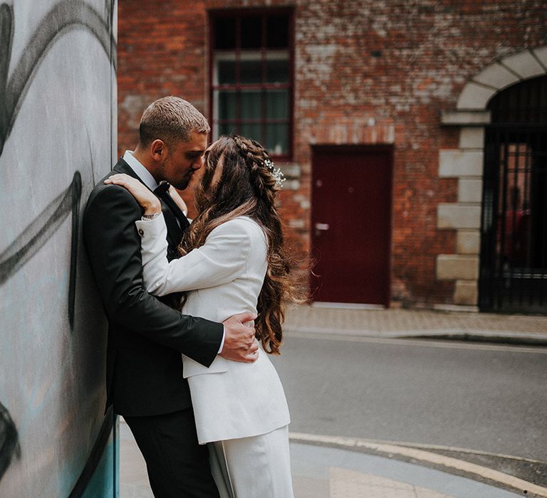 Bride with long wavy half up half down hairstyle in a white suit kissing her groom in a tuxedo 