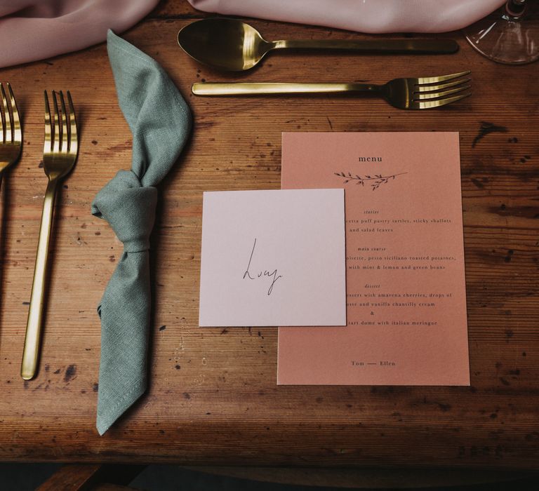 Pink & white stationery on wooden table next to sage green cloth napkin and gold cutlery