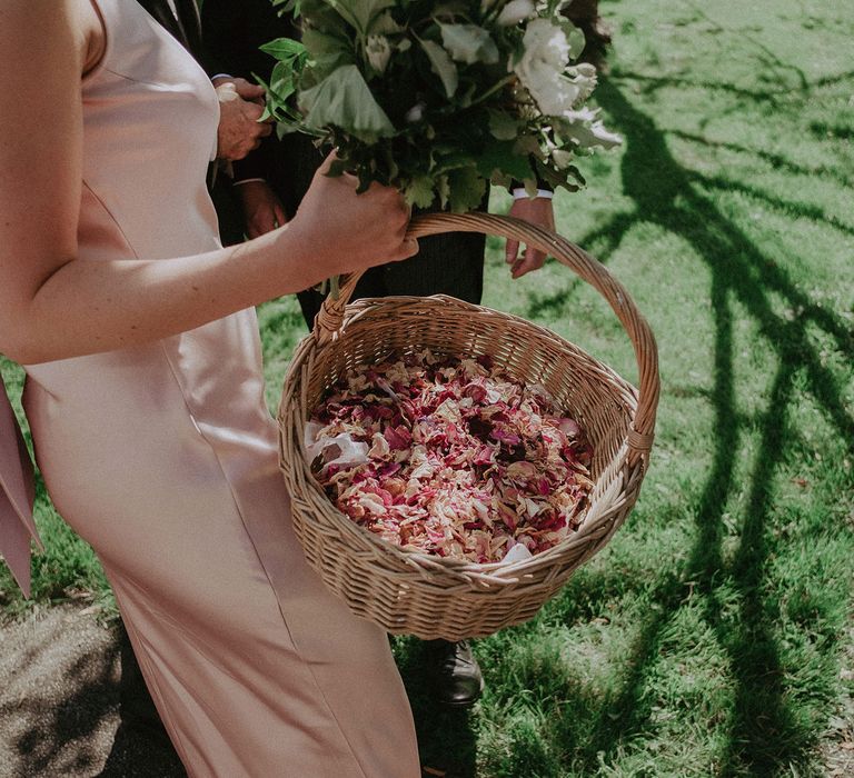 Bridesmaid in pink satin bridesmaid dress holds basket of dried pink rose petals outside church after wedding ceremony in Surrey