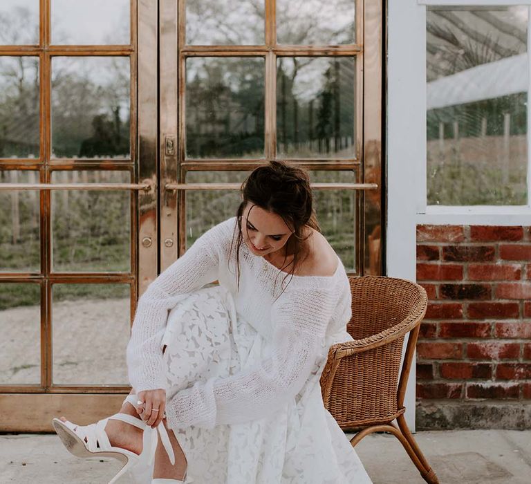 Bride in a lasercut lace skirt and fine knitwear jumper putting on her lace up Emmy London wedding shoes 