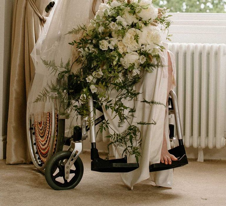 Wedding morning bridal preparations with bride in wheelchair wearing a getting ready robe holding her white and green trailing wedding bouquet 