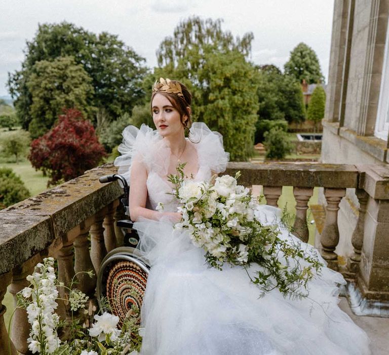 Portrait of the bride in a wheelchair in a tulle wedding dress with gold crown holding a white and green wedding bouquet on the balcony at Bourton Hall 