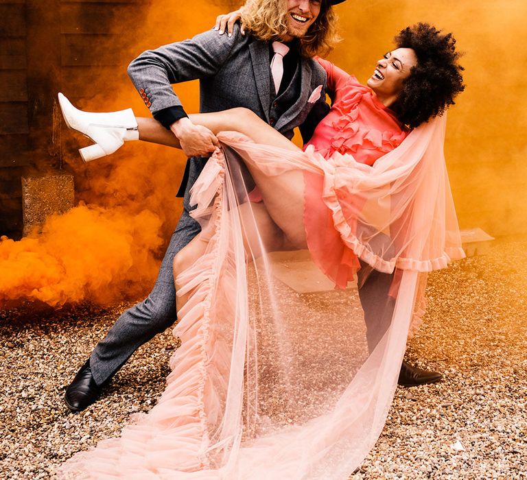 Groom in a grey check suit leaning his bride over in a red dress, white boots and pink veil with an orange smoke bomb going off in the background 