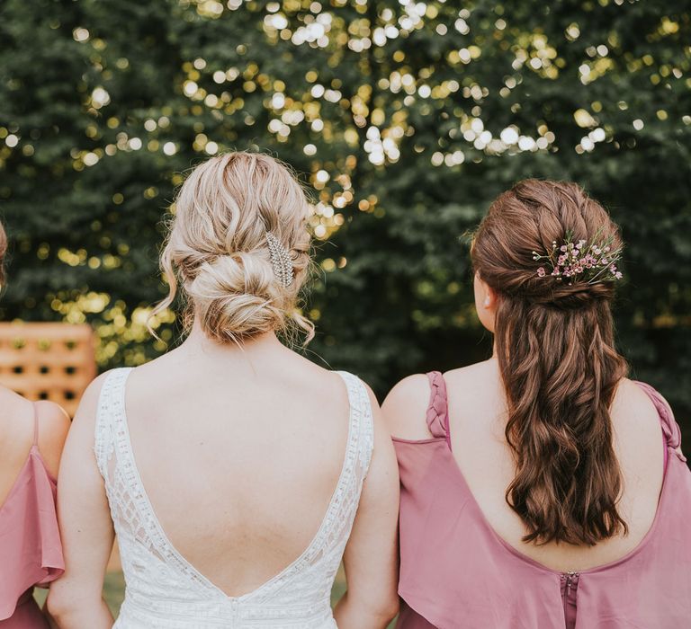 Bride with plaited bun in low v back Justin Alexander wedding dress stands in between bridesmaids with curled hair in dusky pink bridesmaid dresses outside at Tythe Barn wedding with barn wedding flowers