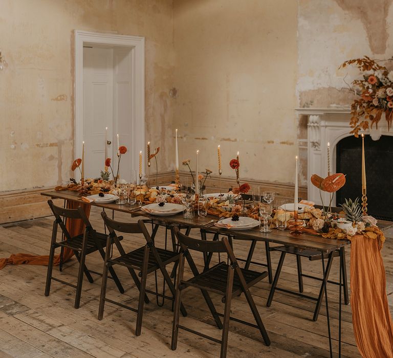 Dark brown wooden table and chairs dresses with an orange table runner, taper candles and dahlia and anthurium flower stems 