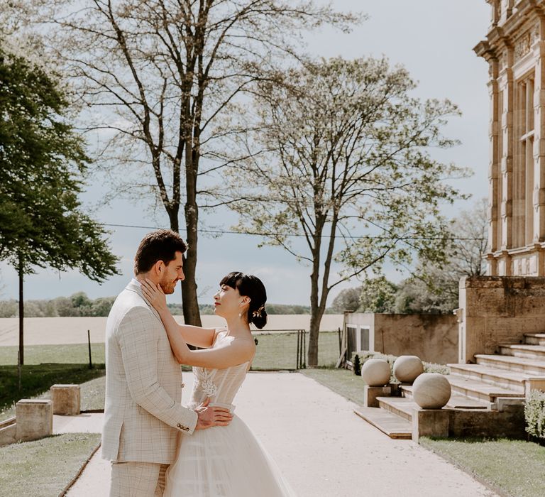 Portrait of the timeless bride with a sleek bridal up do and princess wedding dress with tulle skirt holding her grooms face in the grounds at Bylaugh Hall 