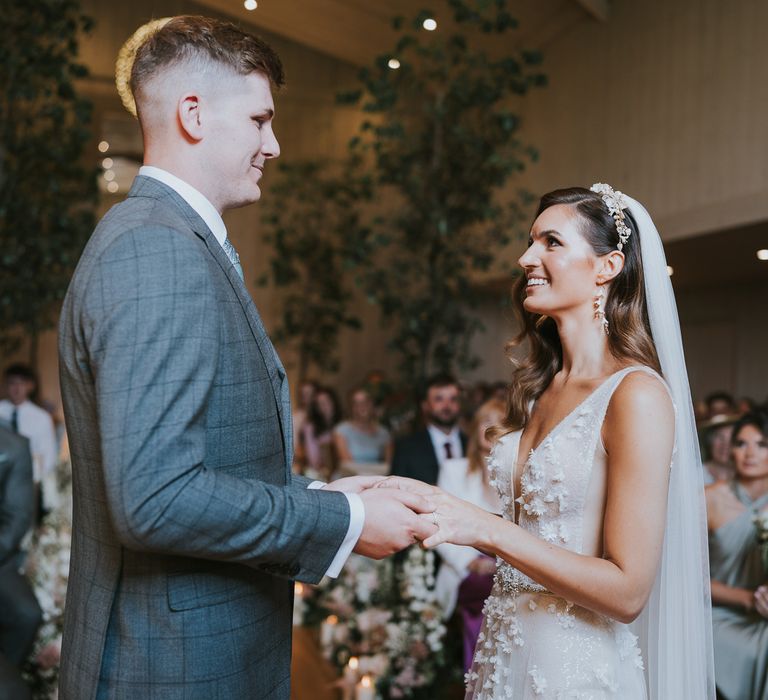 Bride in applique Berta wedding dress, floral headband and veils holds hands with groom in grey checked suit in front of seated wedding guests during wedding ceremony at Primrose Hill Farm