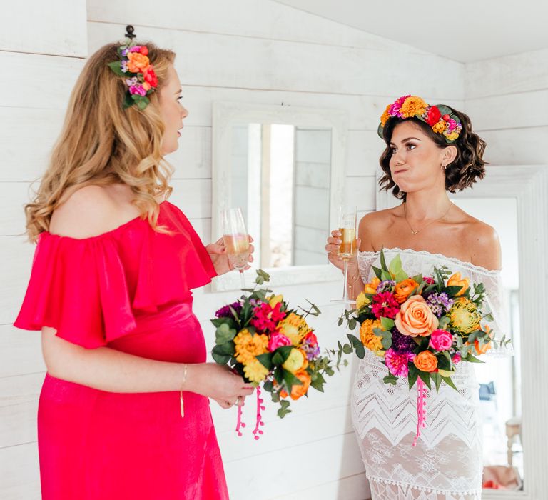 Bride in off the shoulder Grace Loves Lace wedding dress with colour flower crown and colourful bridal bouquet stands with bridesmaid in pink bridesmaid dress as hey both hold glasses of sparkling wine