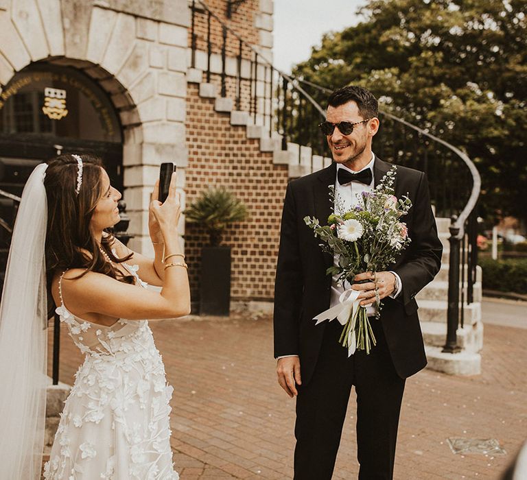 Bride takes photo of the groom who holds her floral bouquet and wears sunglasses 