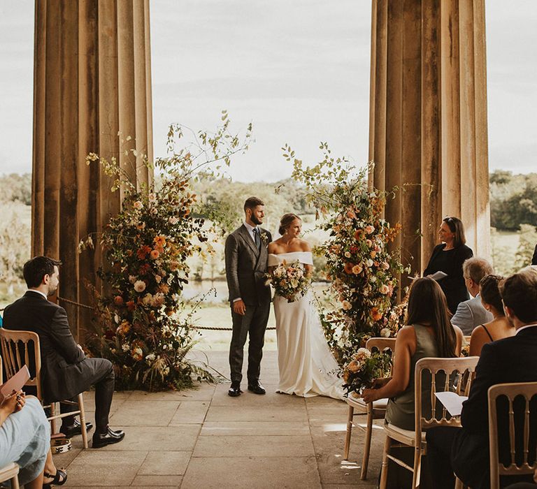 Bride & groom stand in front of floral archway next to large pillars outdoors during wedding ceremony