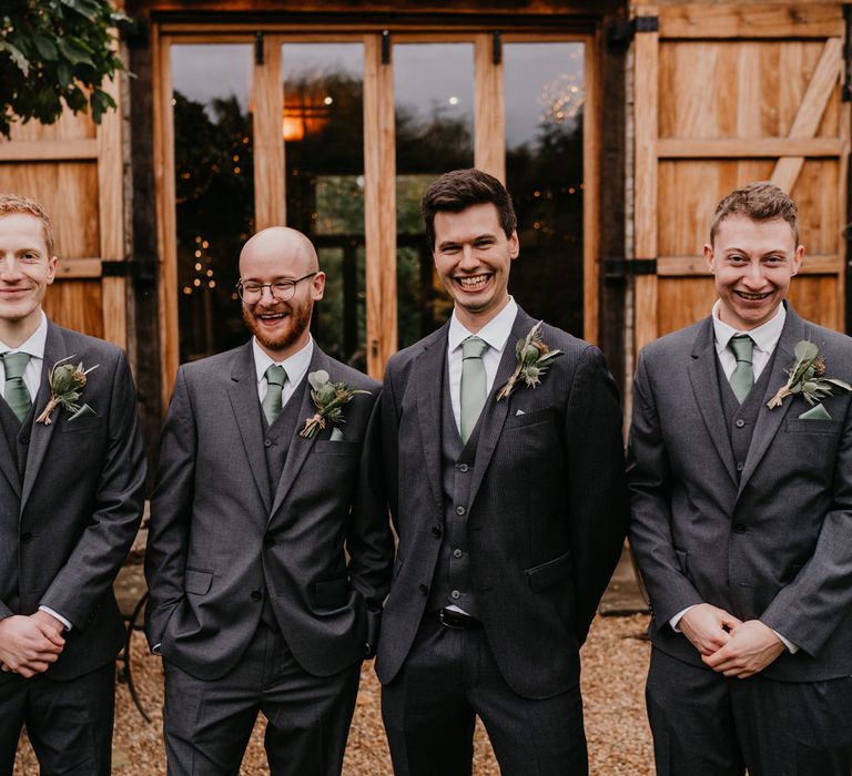 Groom and groomsmen in grey three piece wedding suits, green ties and thistle buttonholes stand outside barn after wedding ceremony