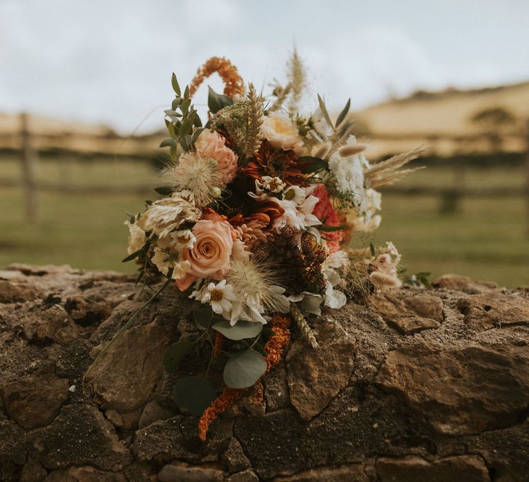 Mixed summer bridal bouquet with dusty pink roses, daisy and various florals sits on stone wall for farm wedding