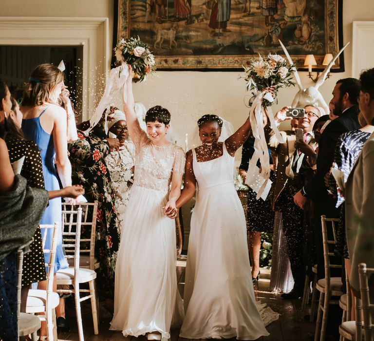 Tow brides walking up the aisle of their Elmore Court wedding ceremony holding their bouquets in the air 